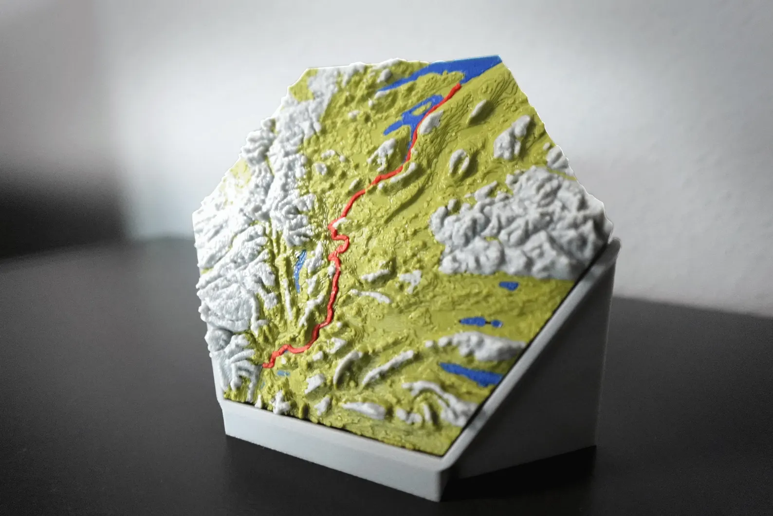 3D-gedrucktes topografisches Modell mit markierter Route in Rot. Detaillierte Landschaft mit Erhebungen, Seen (blau) und Gebirgen (grau). Ideal als personalisiertes Geschenk oder Wanddeko für Wanderer und Naturfreunde. Hexagon-Form mit modernem Sockel.