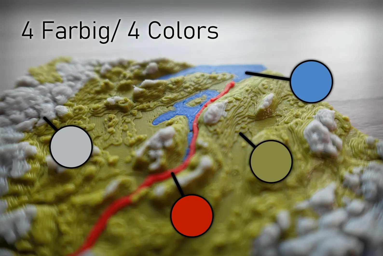 Nahaufnahme eines 3D-gedruckten topografischen Modells mit Beschriftung „4 Farbig/4 Colors“ und farbigen Markierungen: Grau für Berge, Blau für Wasser, Olivgrün für Tiefland und Rot für einen hervorgehobenen Pfad.