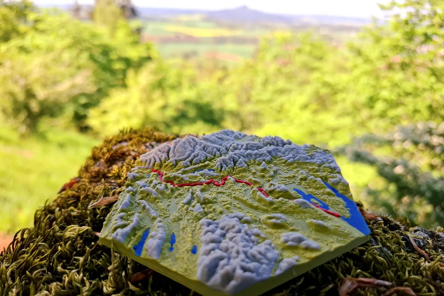 3D-gedrucktes topografisches Modell einer etappe mit farblich hervorgehobener Strecke in Rot, stehendes auf moosbedecktem Baumstumpf in natürlicher, grüner Waldlandschaft mit unscharfem Hintergrund und Aussicht auf Hügel und Felder.