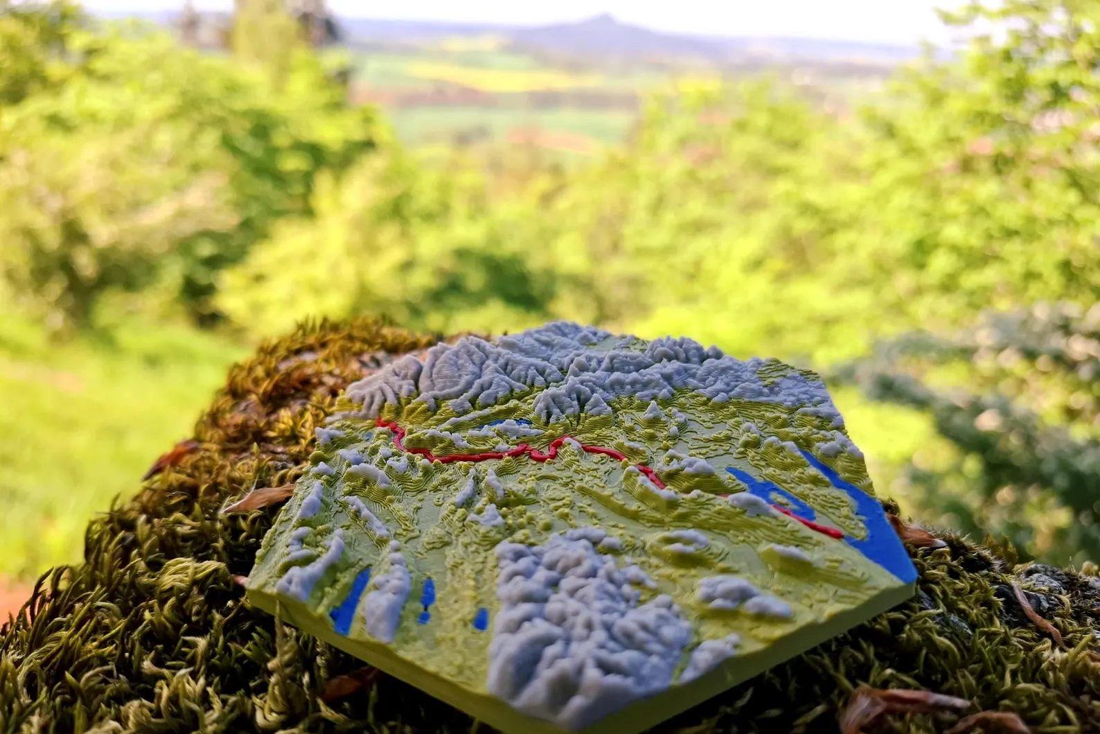 3D-gedrucktes topografisches Modell einer etappe mit farblich hervorgehobener Strecke in Rot, stehendes auf moosbedecktem Baumstumpf in natĂĽrlicher, grĂĽner Waldlandschaft mit unscharfem Hintergrund und Aussicht auf HĂĽgel und Felder.